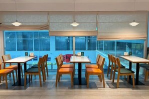 A dining area with tables and chairs, large windows, and hanging lights.