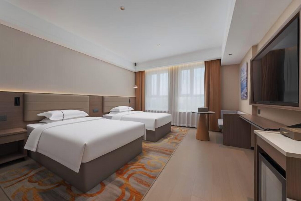 A hotel room with two beds, a TV, a desk, and a window with curtains.
