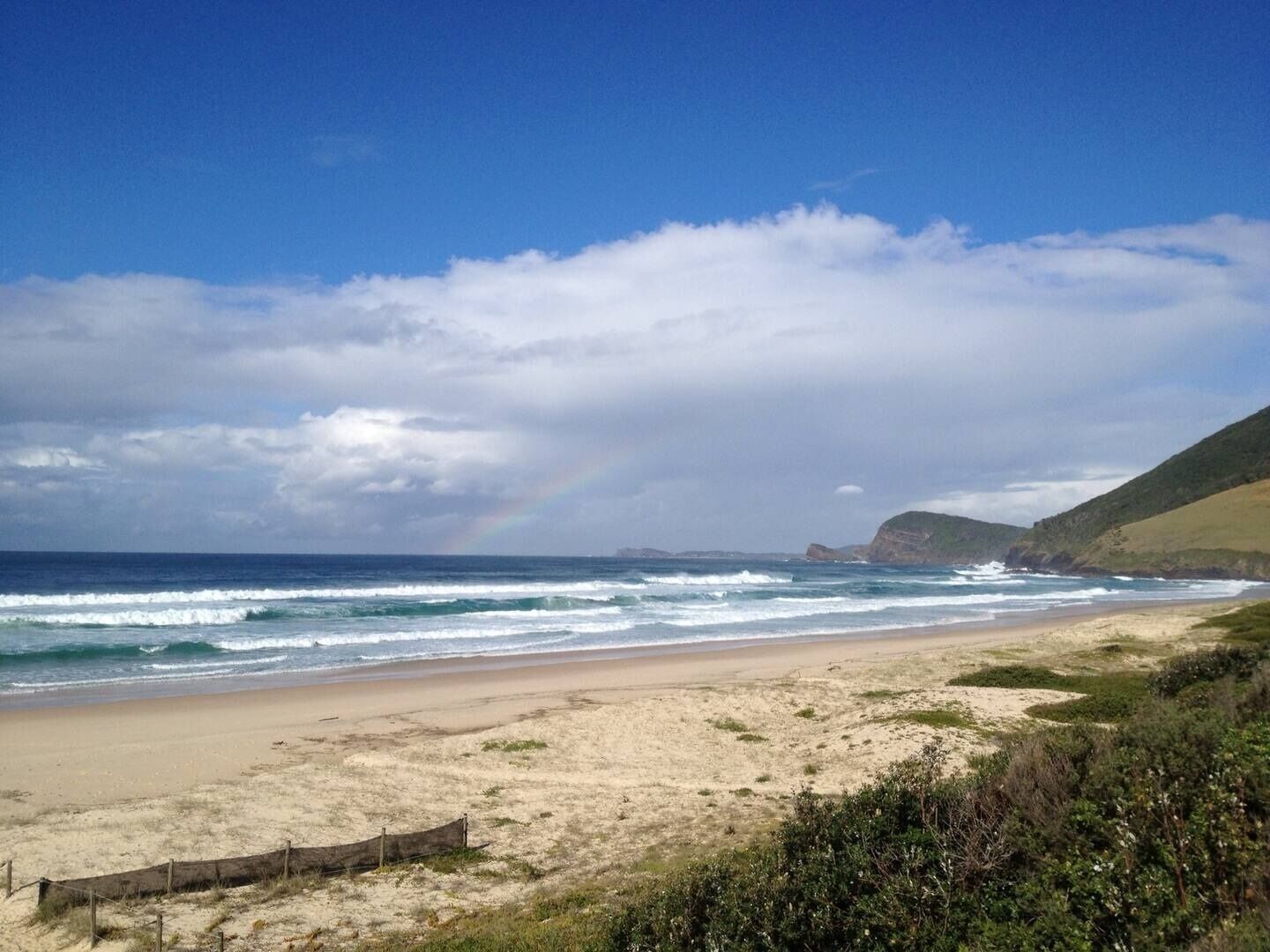 A beach with a fence, waves, and a rainbow.