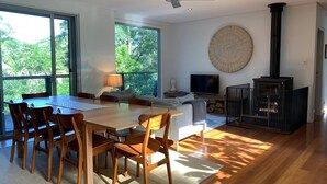 A modern living room with a wooden dining table, chairs, a sofa, a TV, a fireplace, and large windows offering a view of greenery. - Private, Secluded Retreat - Our Reviews Say it All (Smiths Lake)