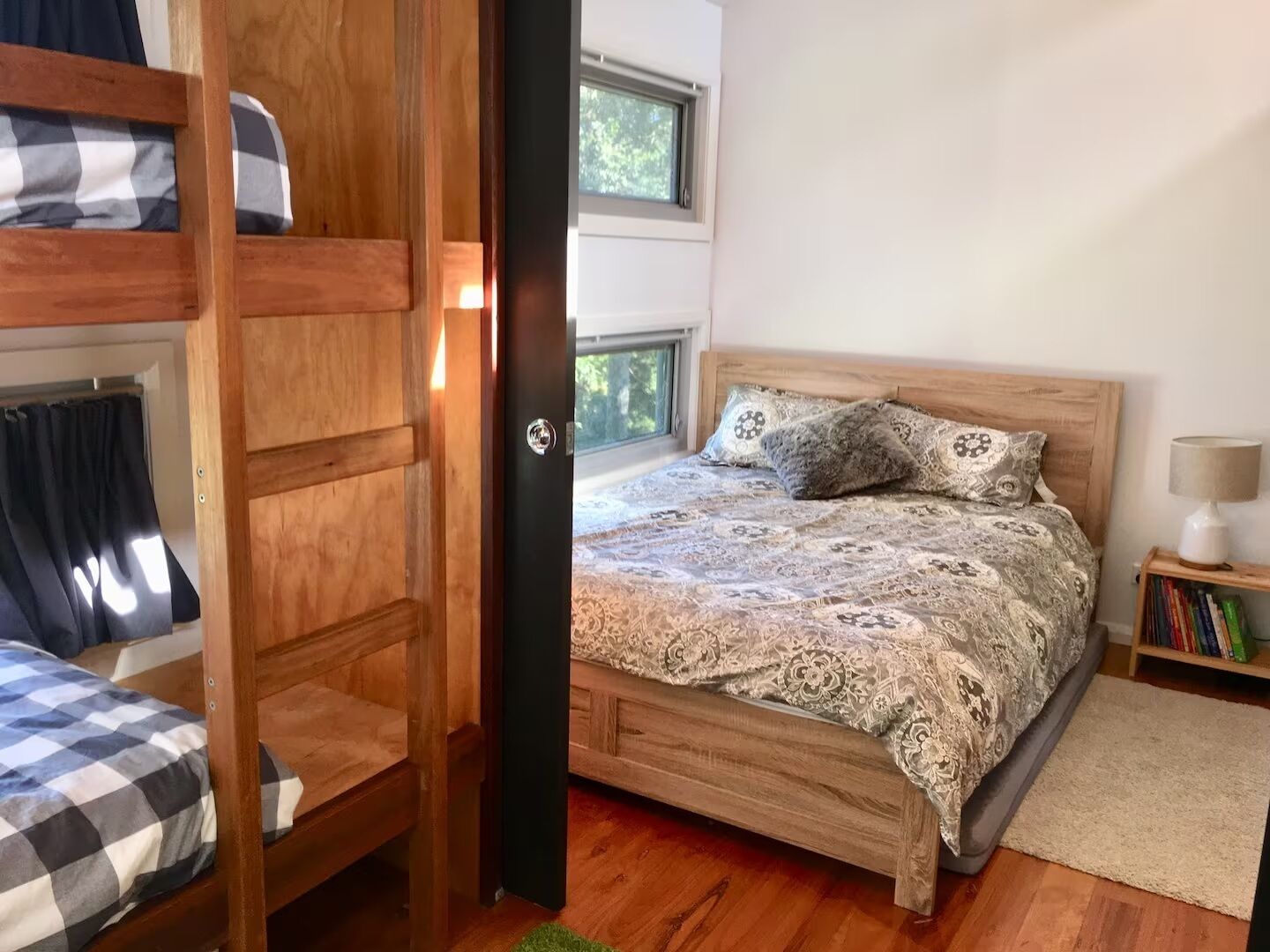 A bedroom with a bunk bed, a single bed with patterned bedding, a bedside table with a lamp, and a bookshelf.