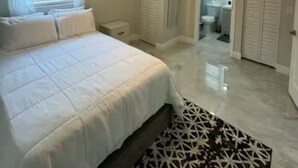 Quarto com cama, ventoinha no teto, mesa de cabeceira e banheiro visível por uma porta aberta.