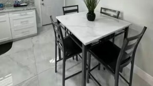 Cozinha moderna com armários brancos, bancada de mármore e área de refeições com mesa branca e cadeiras pretas.