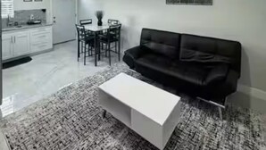 Uma sala de estar moderna com um sofá preto, uma mesa de centro branca e um tapete estampado. Há uma área de jantar com mesa e cadeiras, e uma cozinha com armários e uma janela.