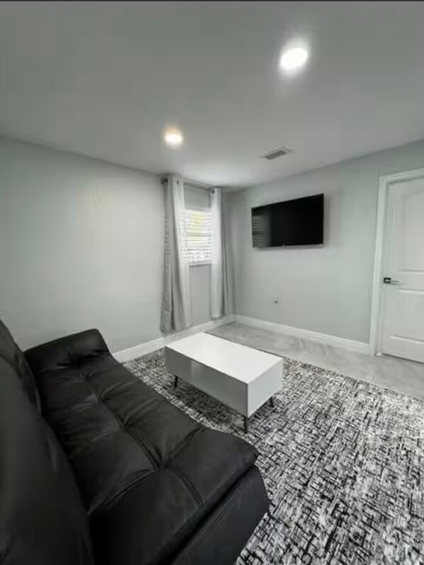A living room with a black leather sofa, a white coffee table, a patterned rug, a flat-screen TV mounted on the wall, and a door.