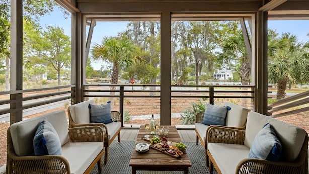 A covered patio with wicker furniture, a wooden table with a fruit platter, and a view of trees and a house.