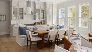 A spacious kitchen with white cabinetry, a wooden dining table, and a seating area with white sofas and wicker chairs.