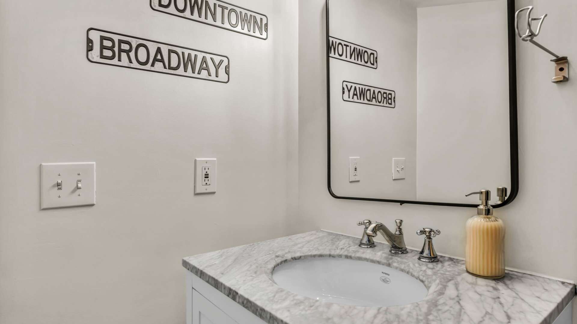 Une salle de bain avec un lavabo en marbre, deux robinets, un distributeur de savon et un miroir avec un cadre noir. On y trouve deux interrupteurs muraux et une prise de courant.