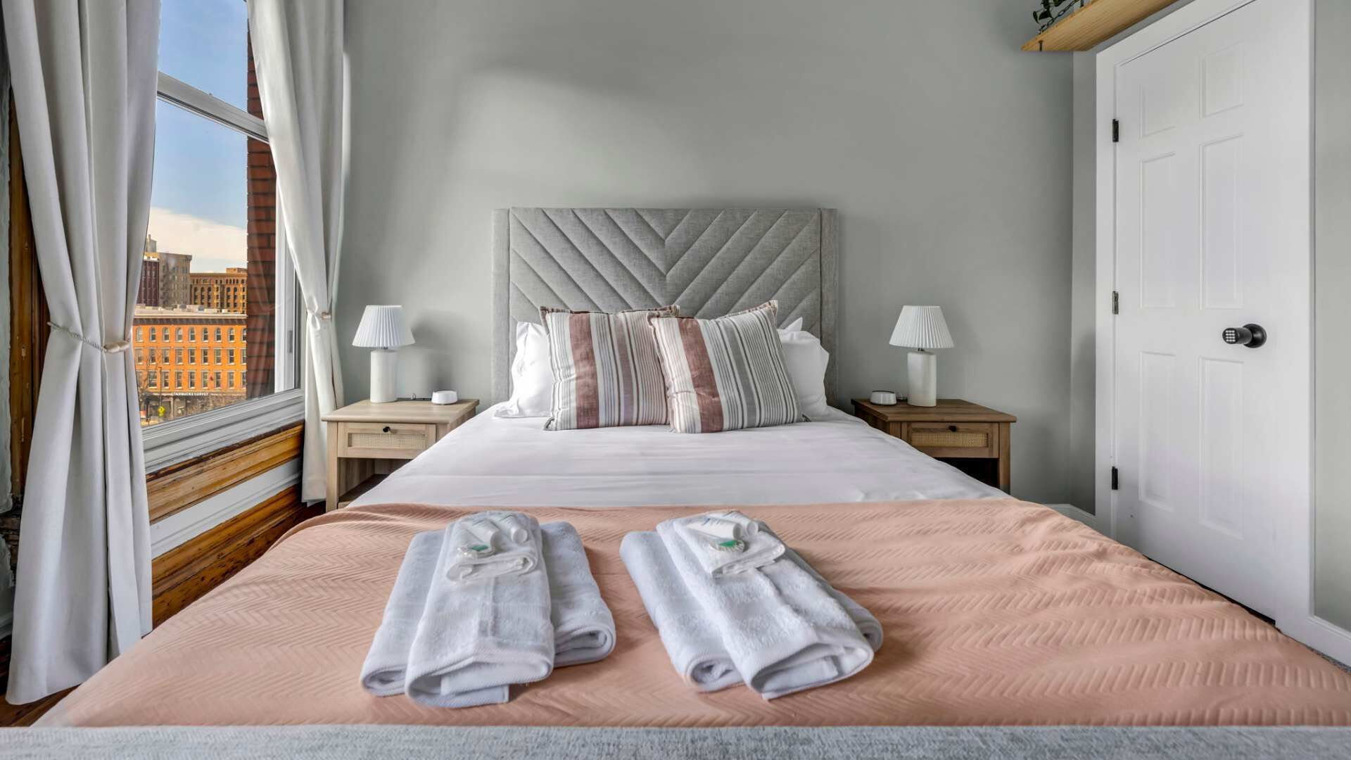 A hotel room with a large bed, two bedside tables with lamps, a view of the city through the window, and a door to the right.