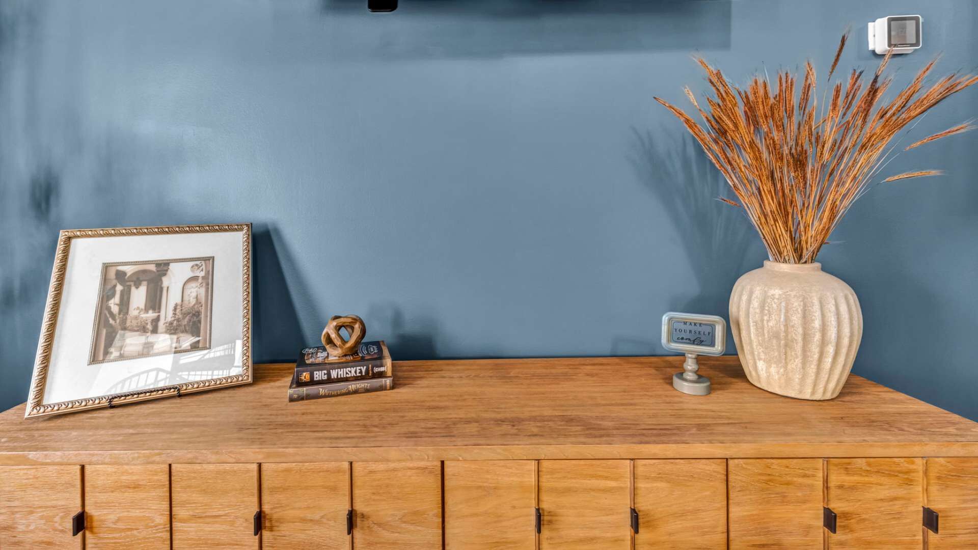 A wooden cabinet with a framed picture, a whiskey bottle, a salt shaker, a vase with dried plants, and a motivational sign.