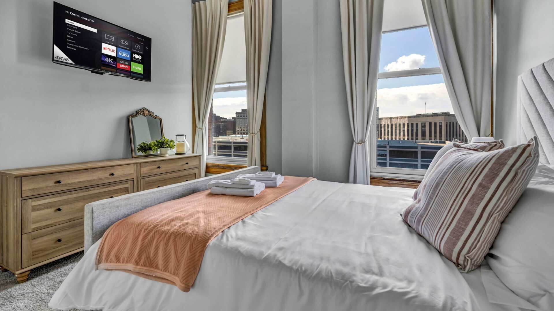 A hotel room with a bed, a dresser, a TV mounted on the wall, and a window with curtains.