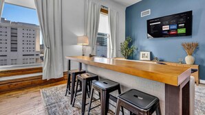 Et moderne hotelværelse med en træbar, to barstole, et TV monteret på væggen og udsigt over byen gennem store vinduer.