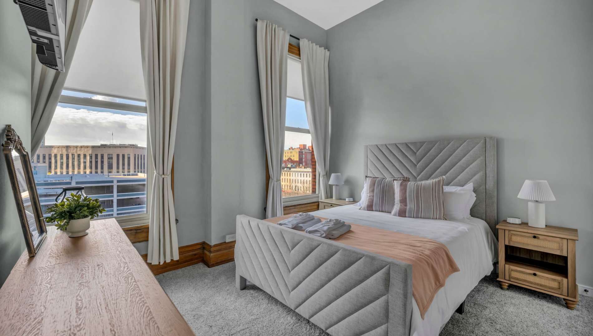 Une chambre d’hôtel moderne avec un grand lit, une table de chevet, une commode et une vue sur le paysage urbain.