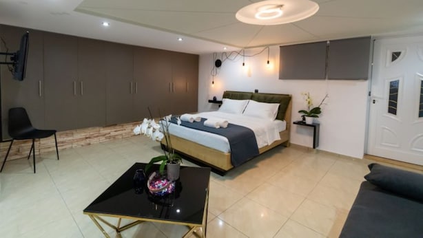 A modern bedroom with a bed, a small table with a vase, a chair, and a sofa. - Black suites (Chania)