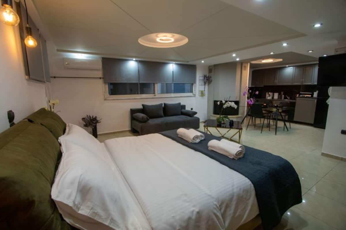 A modern hotel room with a large bed, a sofa, a dining area, and a kitchen.
