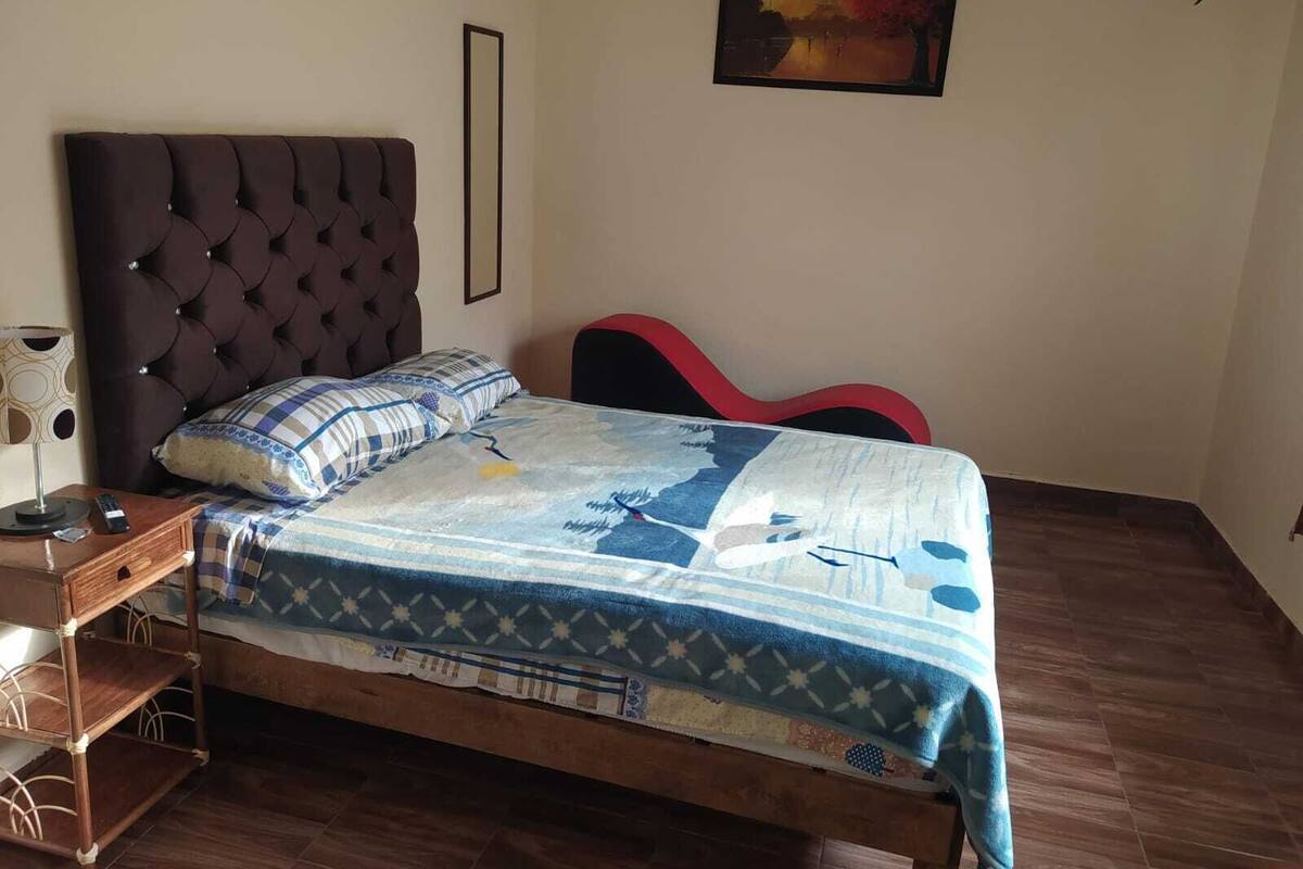 Un dormitorio con cabecera acolchada, cama con colcha estampada, mesita de noche con lámpara y una silla roja.