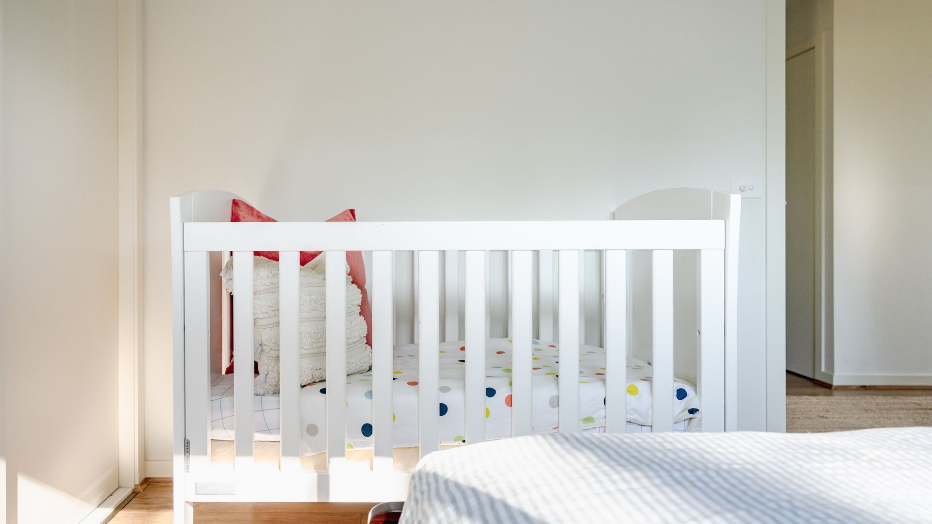 A white crib with a polka dot mattress, a white wall, and a wooden floor.