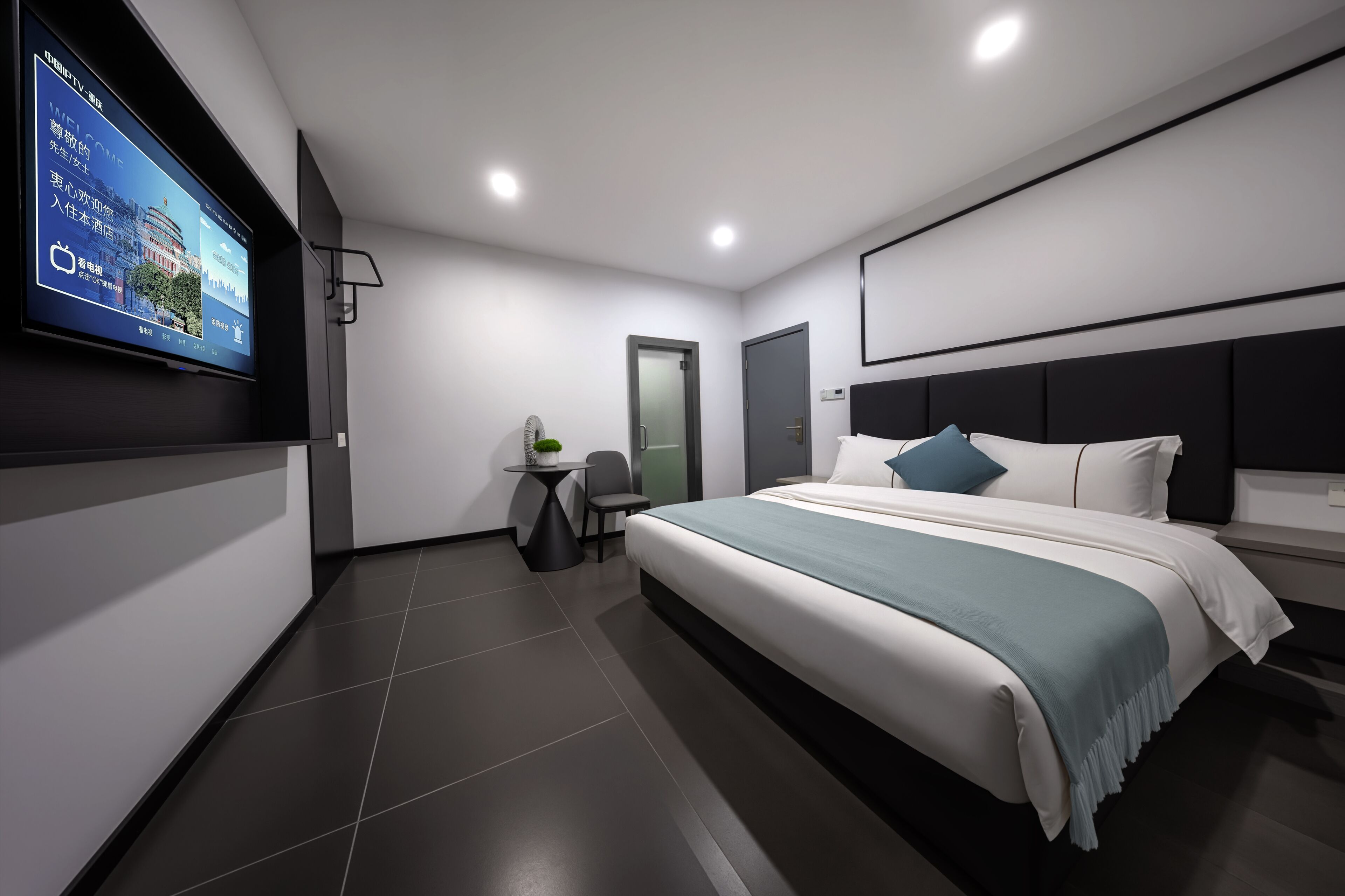 A modern hotel room with a large bed, a flat-screen TV mounted on the wall, a small table with a chair, and a door.