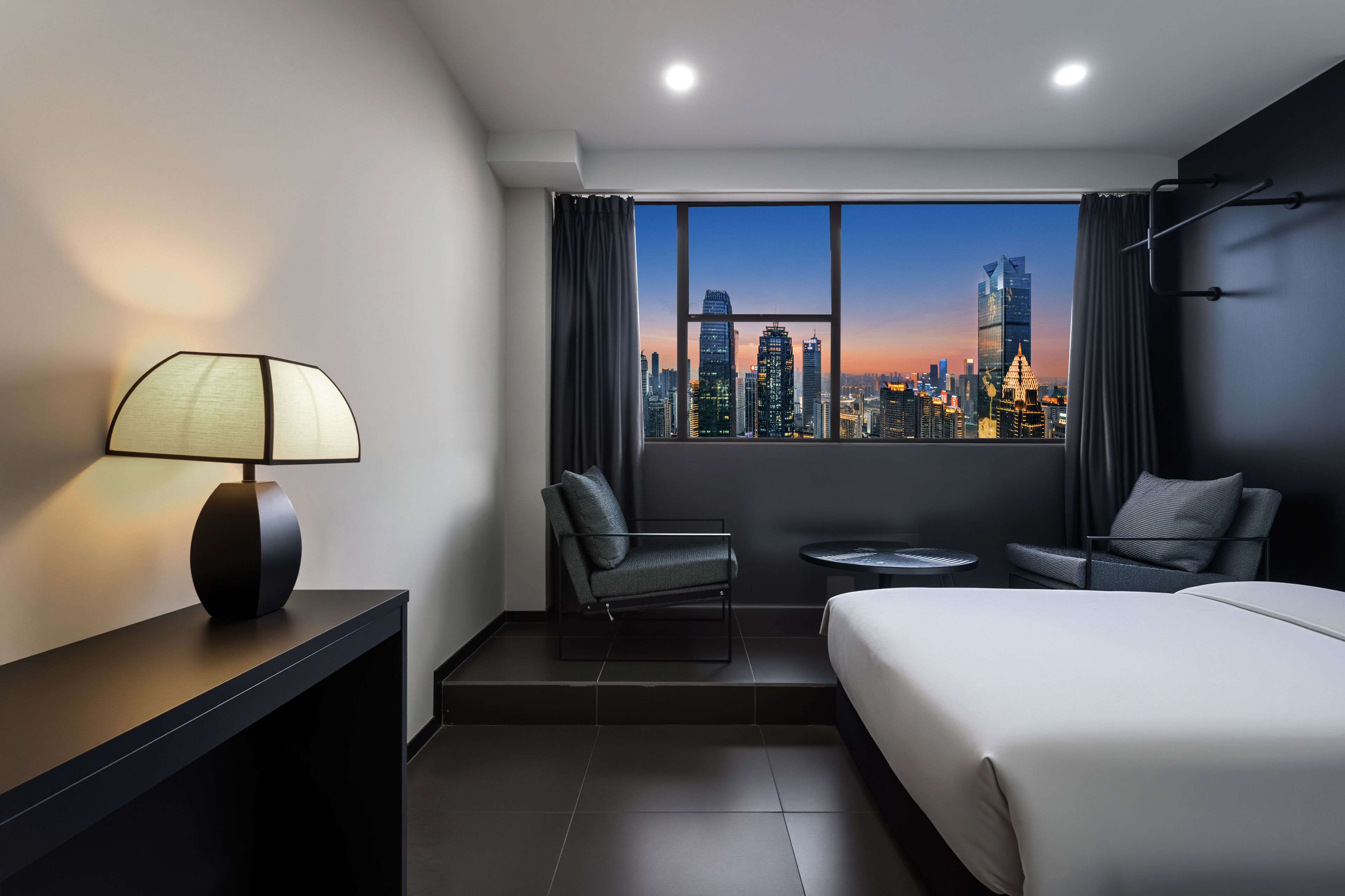 A modern hotel room with a city skyline view, a bed, two armchairs, a small table, and a lamp.