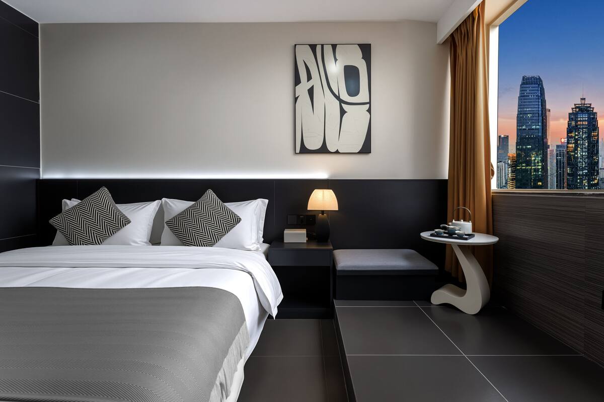Habitación de hotel moderna con una cama grande, una mesita de noche con lámpara, una pequeña mesa con juego de té y vista a un paisaje urbano por la ventana.