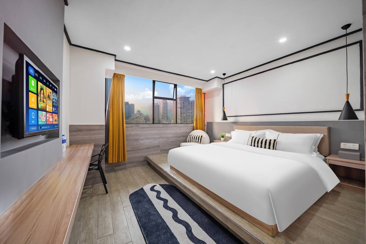 Una habitación de hotel moderna con una cama grande, un televisor en la pared, vista a la ciudad y piso de madera.