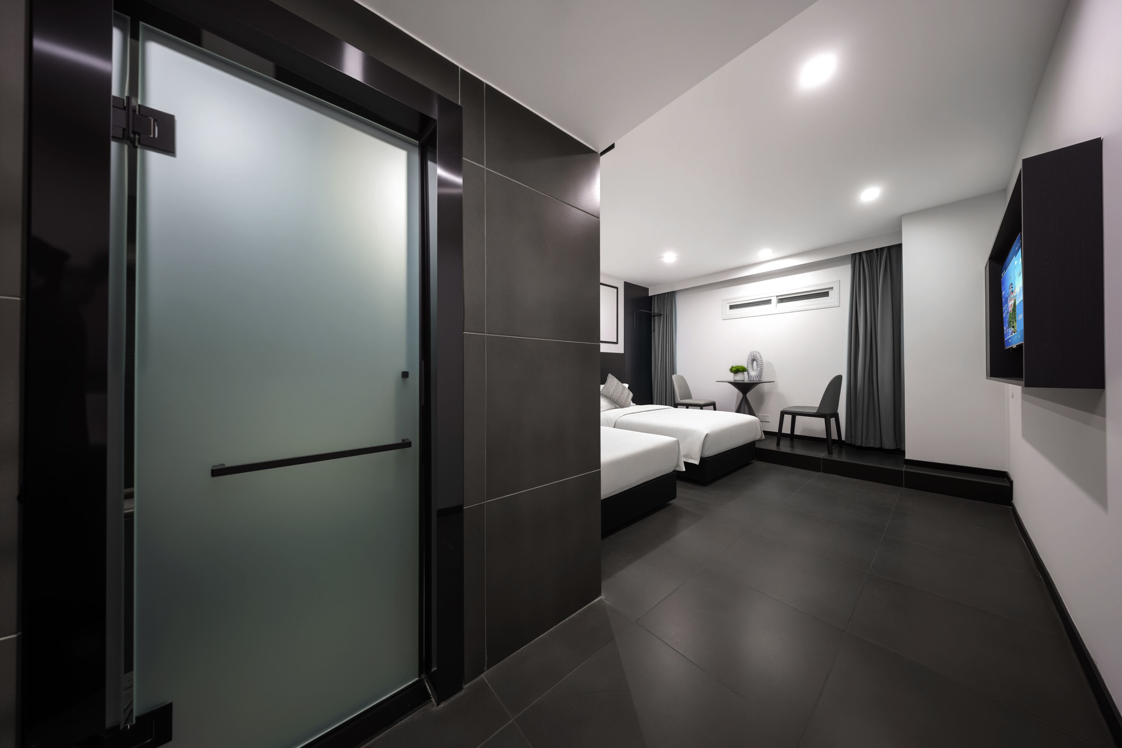 Una habitación de hotel moderna con puerta corredera, cama, mesita de noche, sillón y televisor montado en la pared.