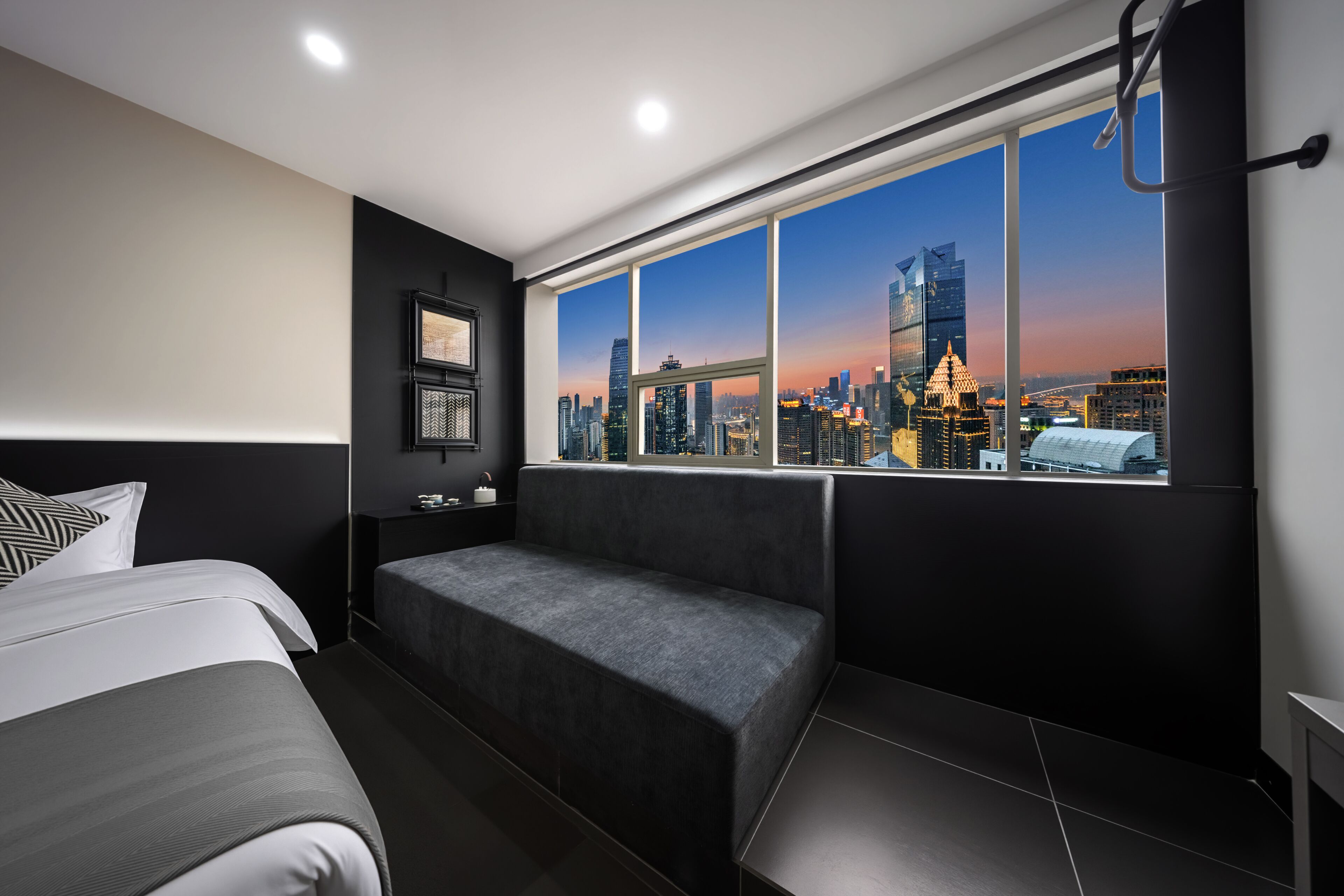A modern hotel room with a large window, a bed, a sofa, and a nightstand.
