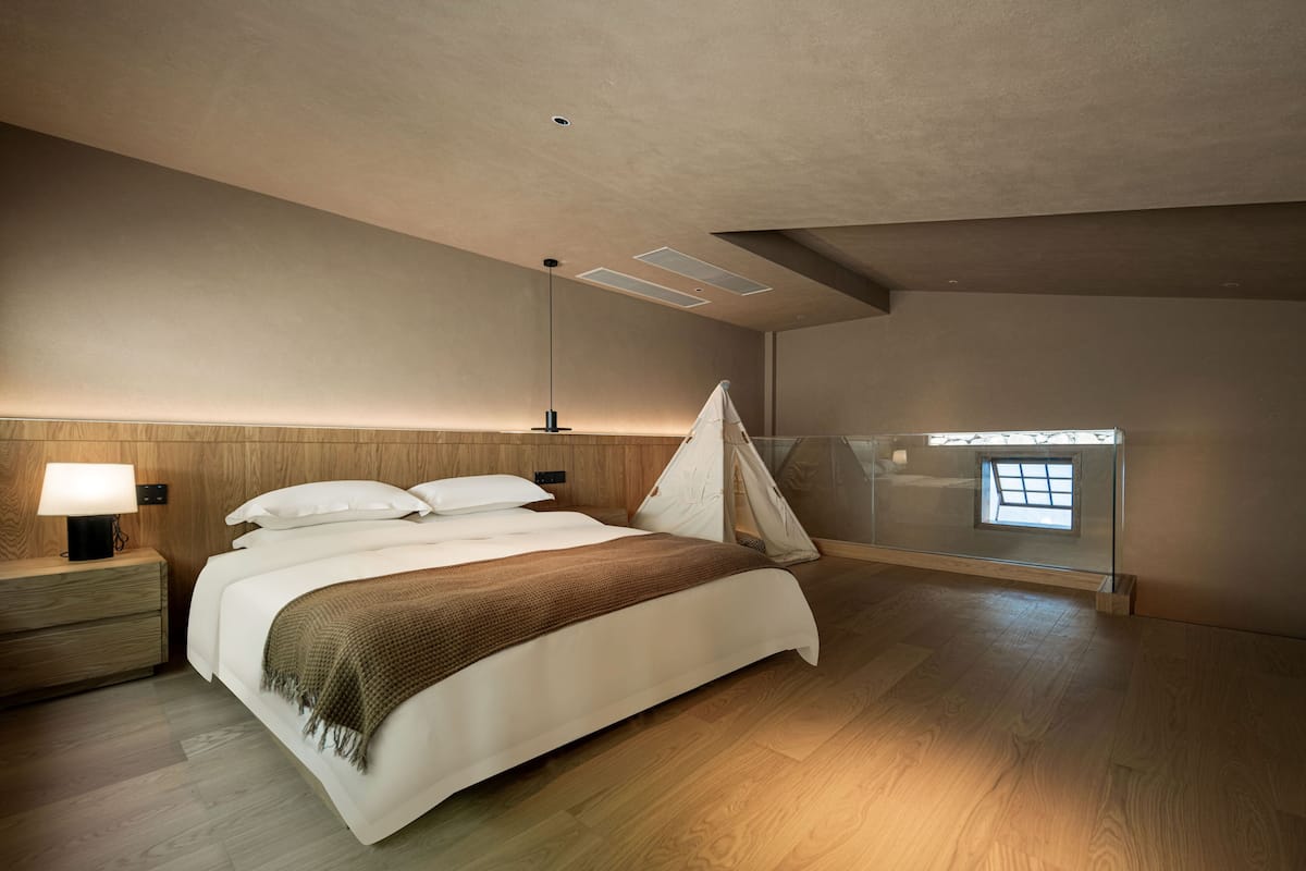 A modern bedroom with a large bed, wooden headboard, bedside lamp, and a tent-like structure against the wall.