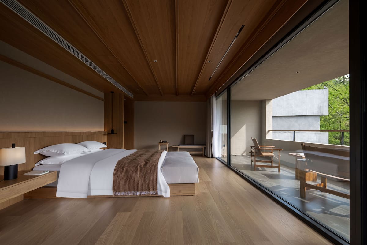 A modern bedroom with a large bed, bedside tables, a bench, and a view through large windows.