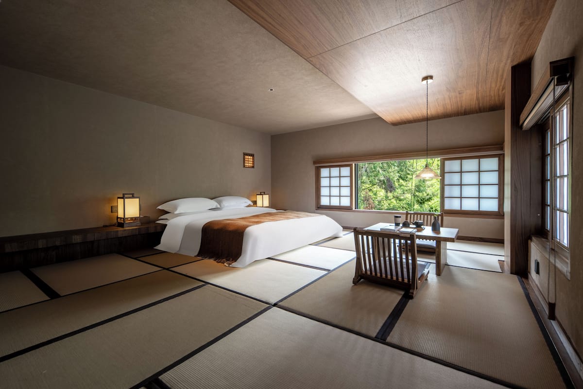 A traditional Japanese-style room with tatami flooring, a bed with white bedding, a small table with a chair, and a window with a view of greenery.
