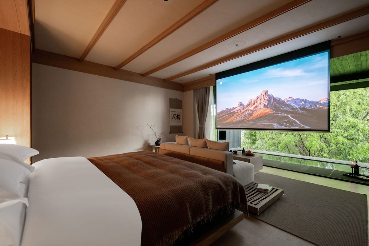 A modern hotel room with a large bed, a sofa, a TV screen displaying a mountain landscape, and a balcony with a view of trees.