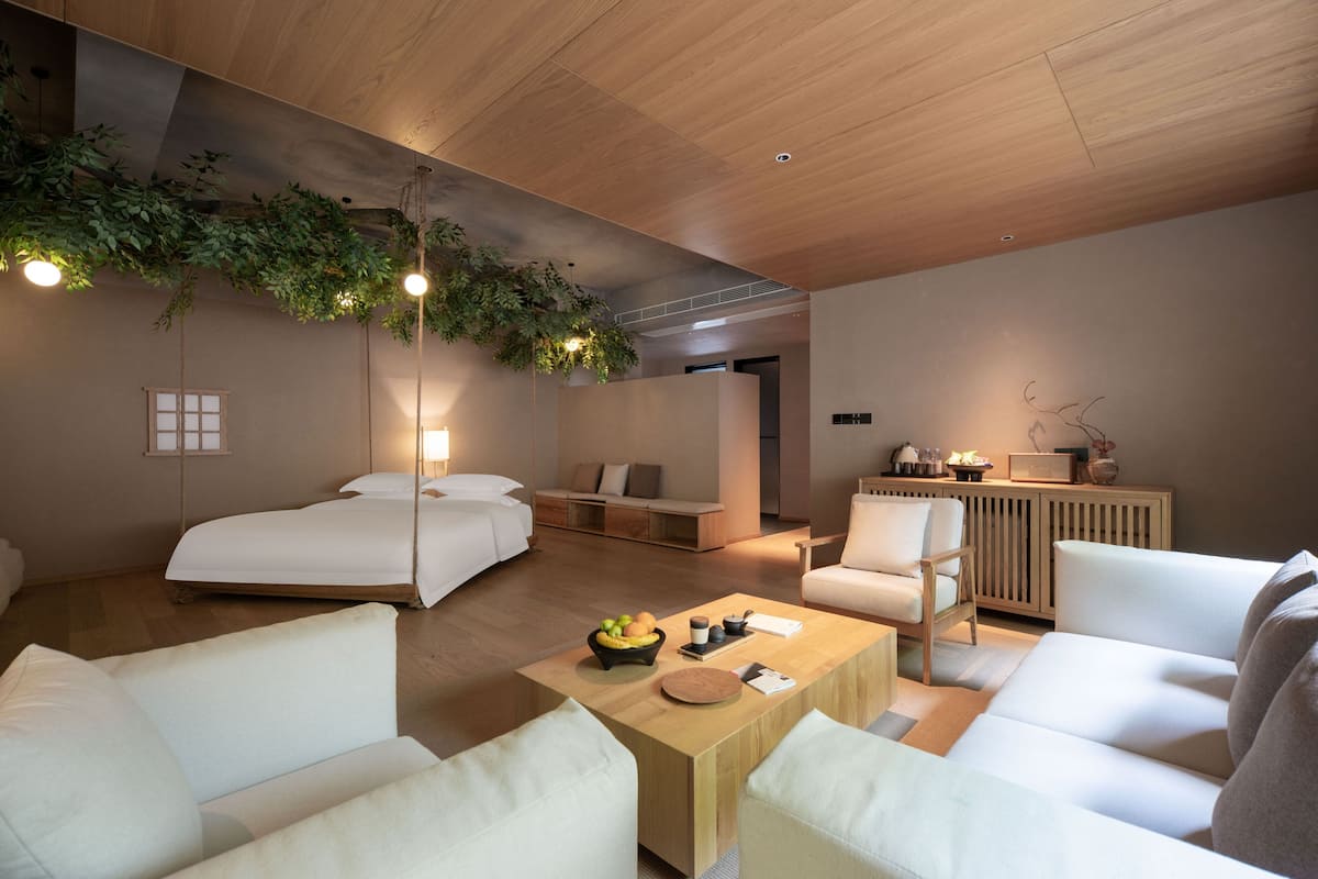 A modern hotel room with a wooden ceiling, a large bed, a sofa, a coffee table with fruits, and a small kitchenette area.