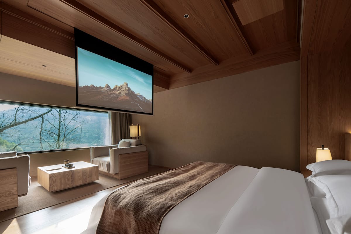 A modern hotel room with a large bed, a TV mounted on the wall, a sofa, and a bedside table.