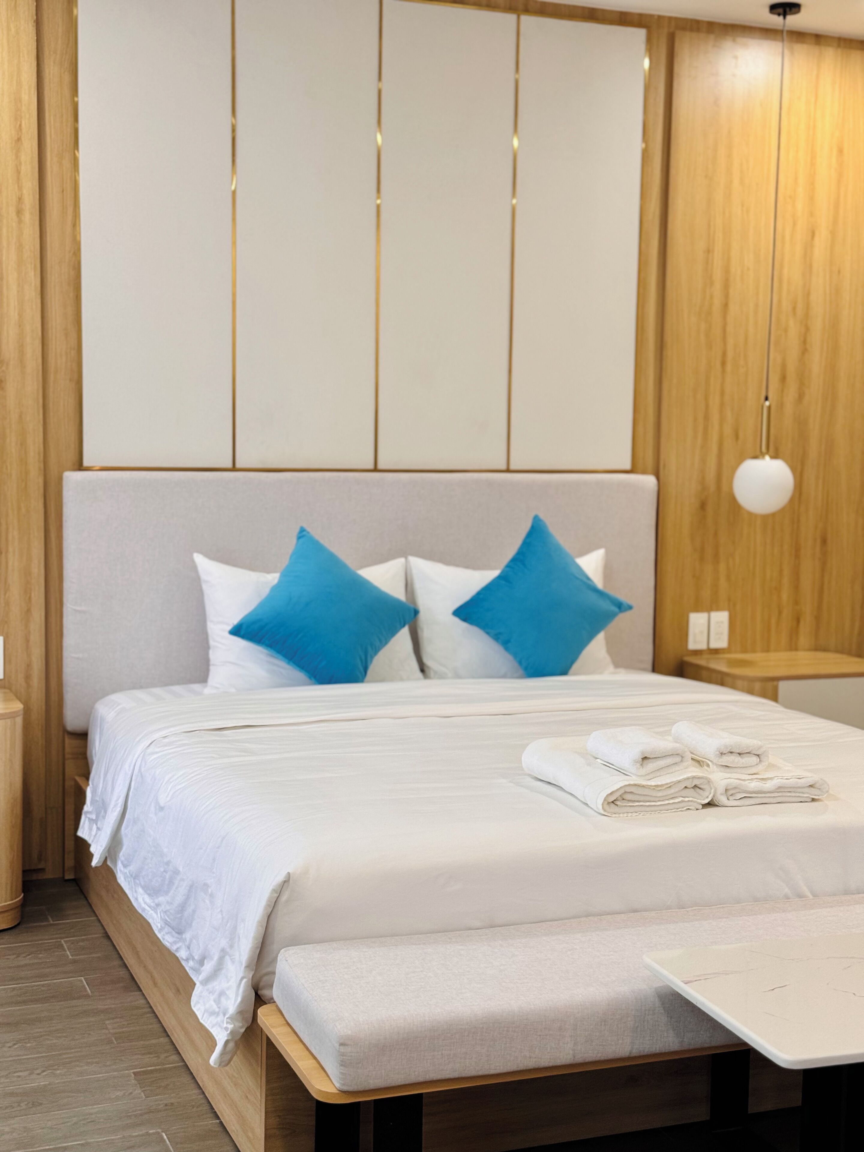 A neatly made bed with white linens and blue pillows, a bedside table with a lamp, and a wooden headboard.