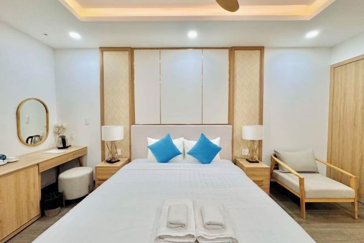 A modern hotel room with a large bed, a wooden desk, a chair, and a wardrobe.