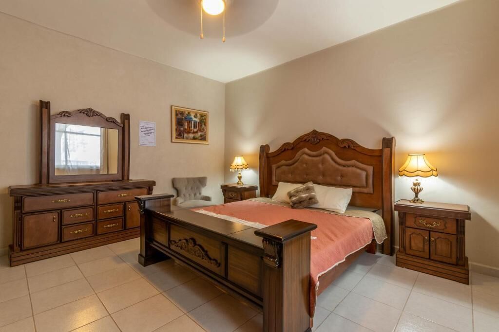 Un dormitorio con una cama de madera, un aparador, dos mesitas de noche con lámparas, una silla y un espejo.