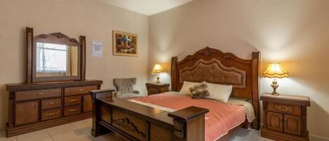 Un dormitorio con una cama de madera, un aparador, dos mesitas de noche con lámparas, una silla y un espejo.
