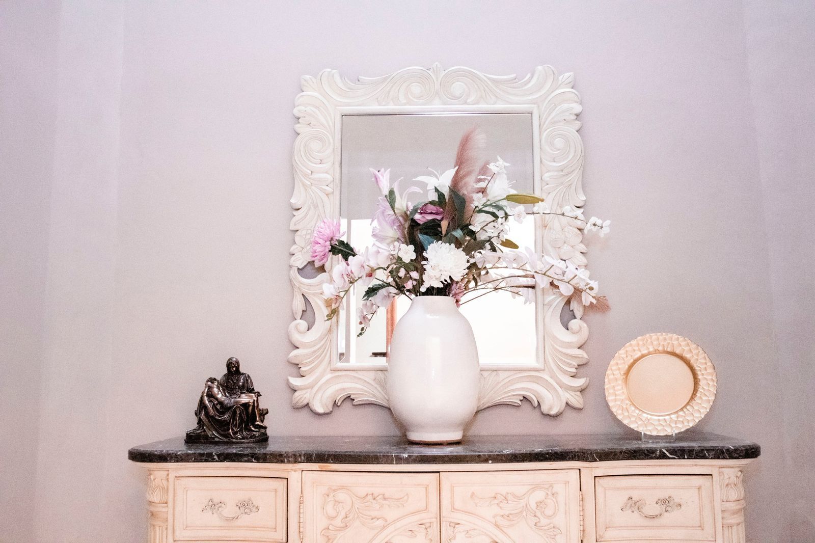 A vanity with a mirror, a vase of flowers, a statue, and a decorative plate.