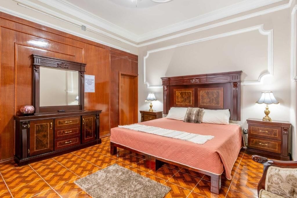 Quarto com cama grande, cabeceira de madeira, duas mesas de cabeceira com luminárias, um espelho amplo e piso com padrão herringbone.