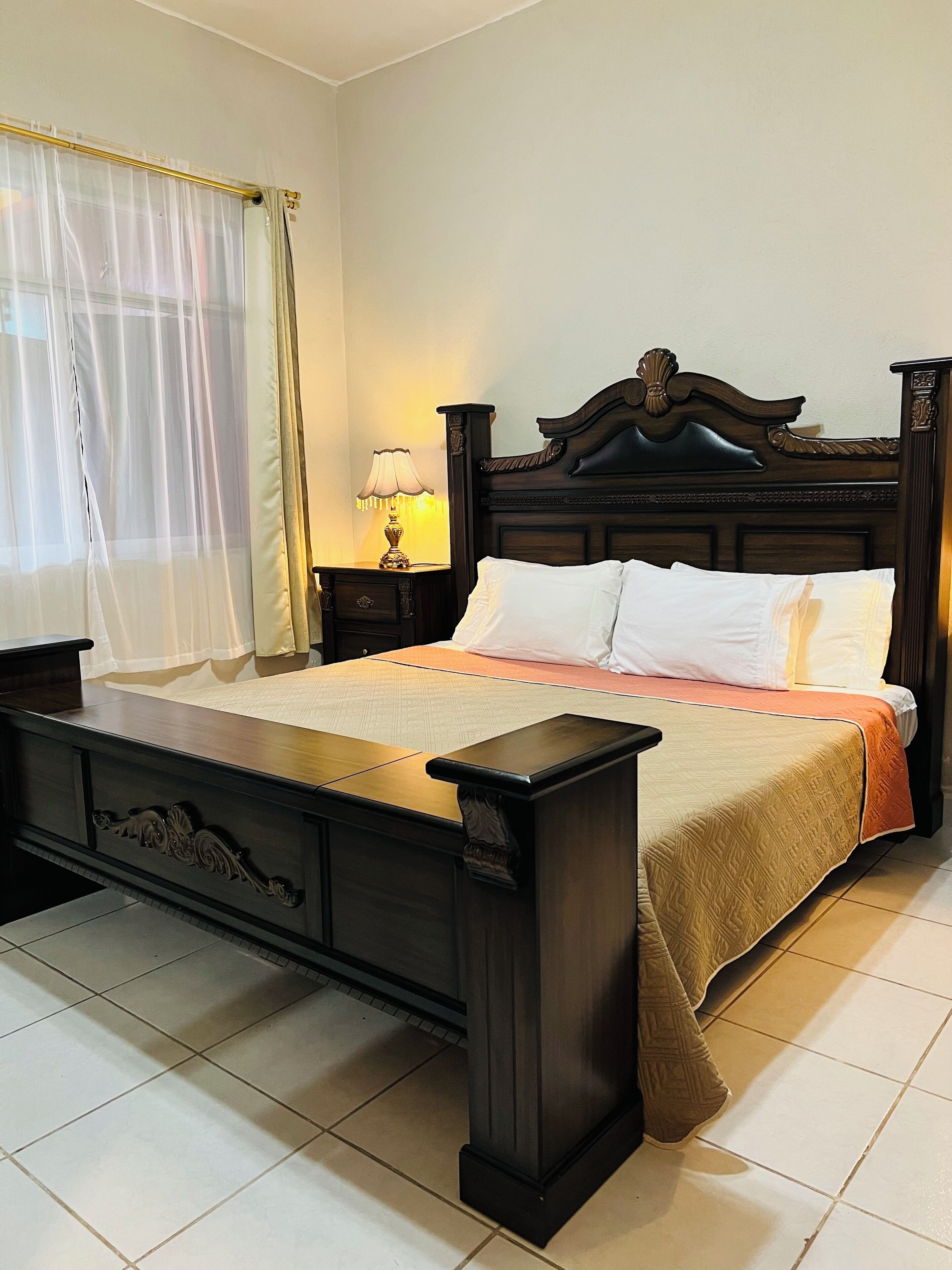 A bedroom with a wooden bed, a nightstand with a lamp, and a window with sheer curtains.