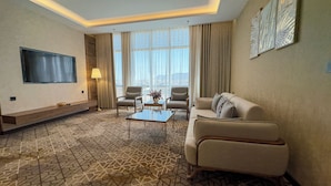 Een hotelkamer met een bank, twee fauteuils, een salontafel, een flatscreen televisie en een groot raam met gordijnen.