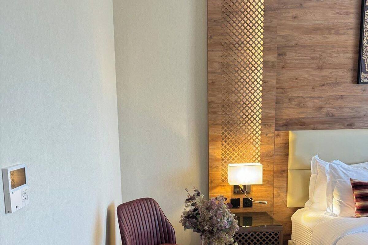 A hotel room with a bed, two chairs, a small table with a vase, a nightstand with a lamp, and a decorative wall panel.
