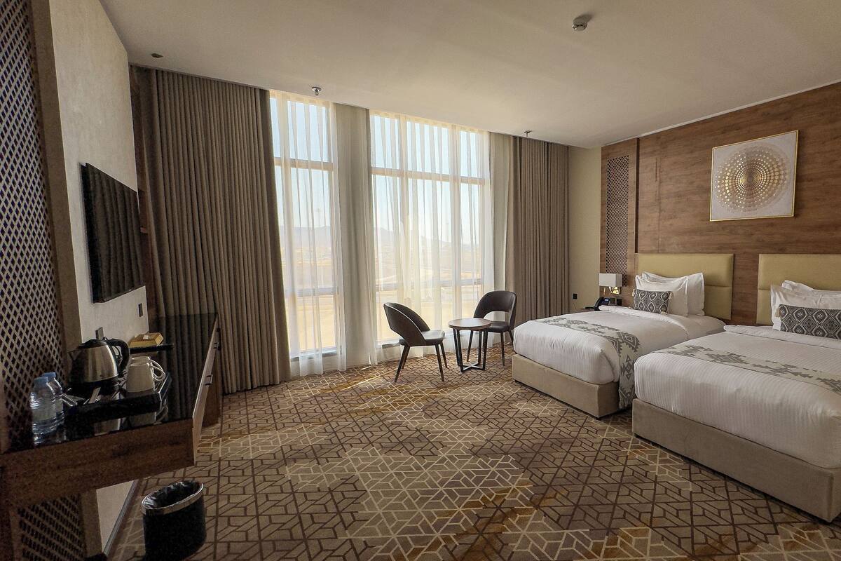 A hotel room with two beds, a desk with a kettle, a chair, and a large window with curtains.