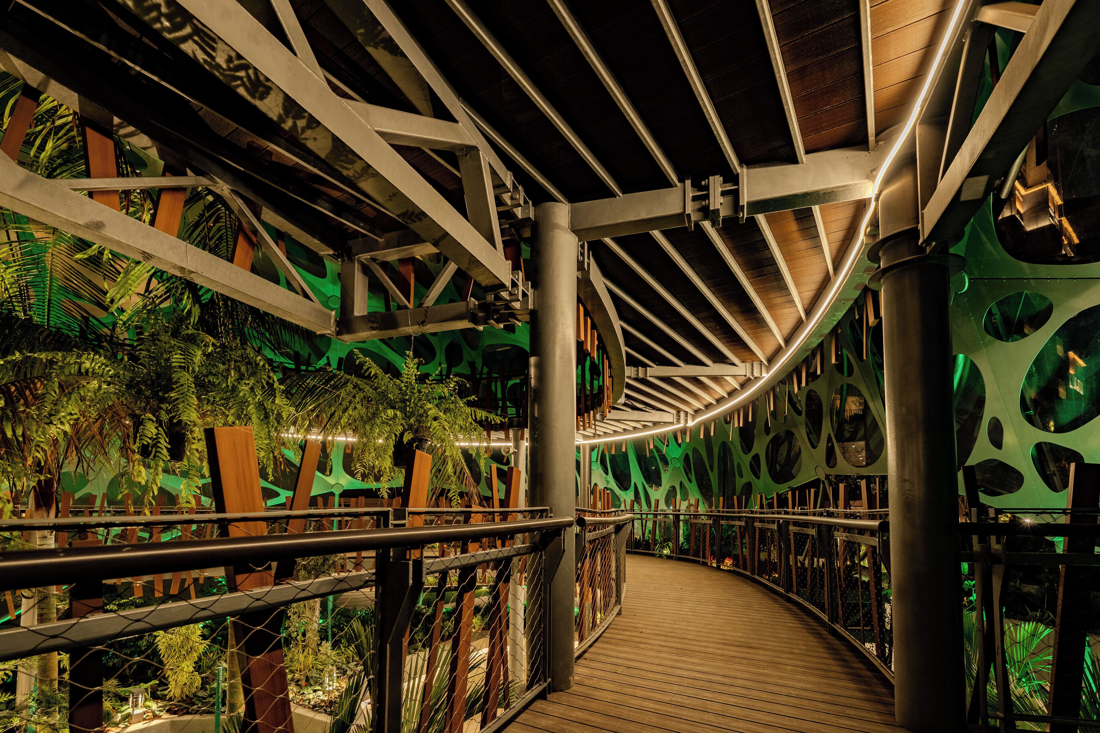 Une passerelle en bois, agrémentée de rampes métalliques, entourée d’une végétation luxuriante et ponctuée d’éléments architecturaux modernes.