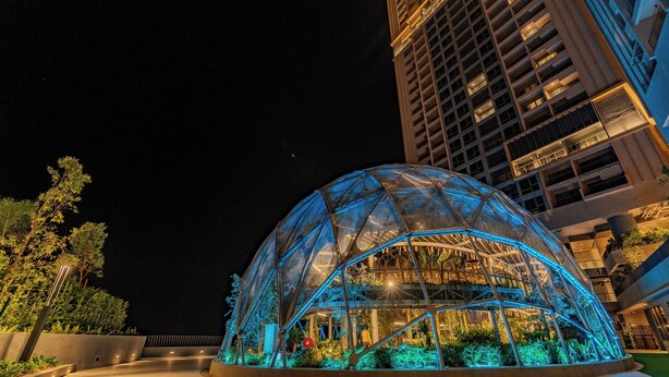 A modern glass structure with a dome roof, surrounded by greenery and illuminated by blue lighting.