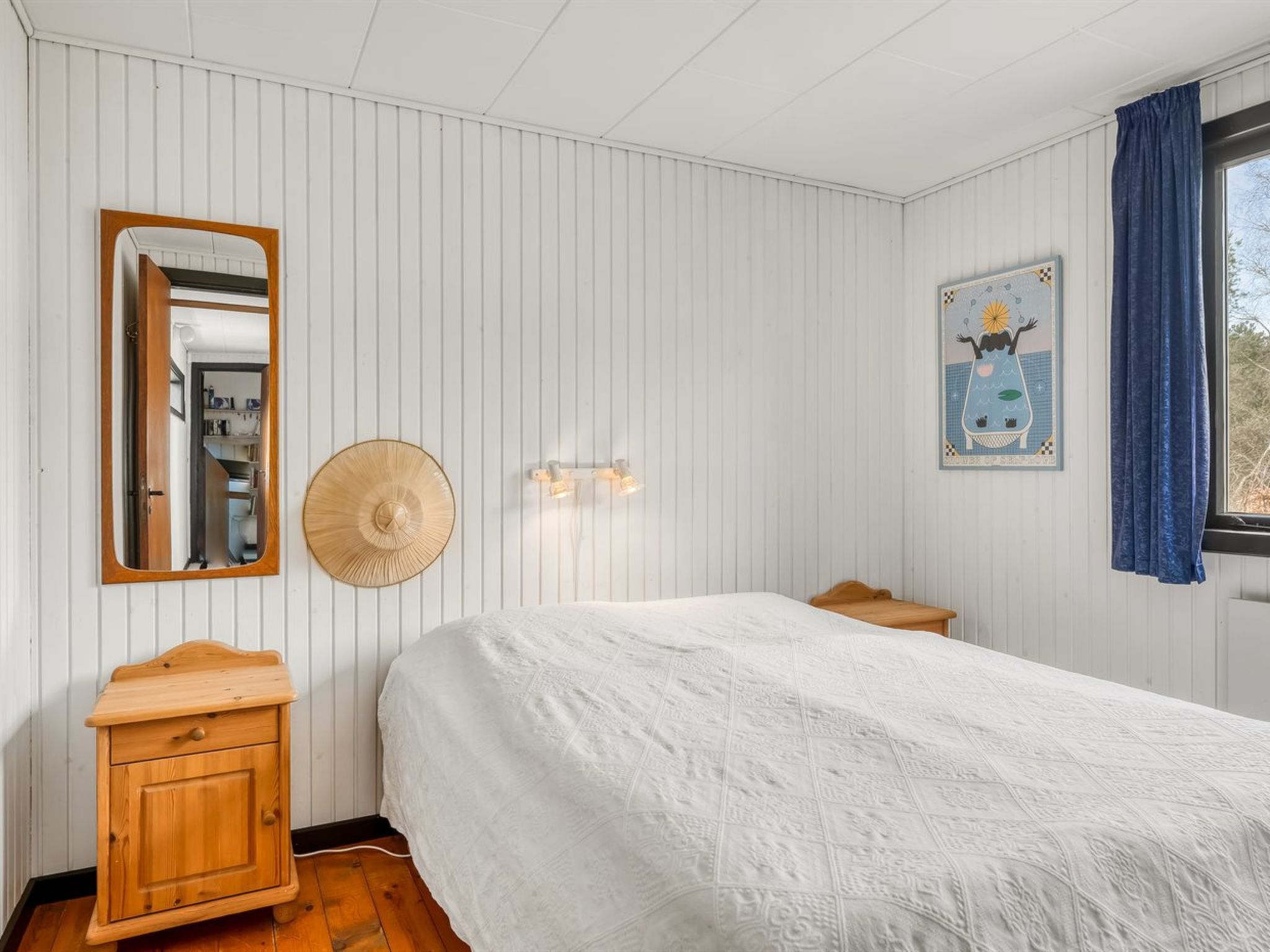 Ein Schlafzimmer mit einem Bett, einem Nachttisch aus Holz, einem Spiegel, einer Wandleuchte und einem Fenster mit Vorhängen.