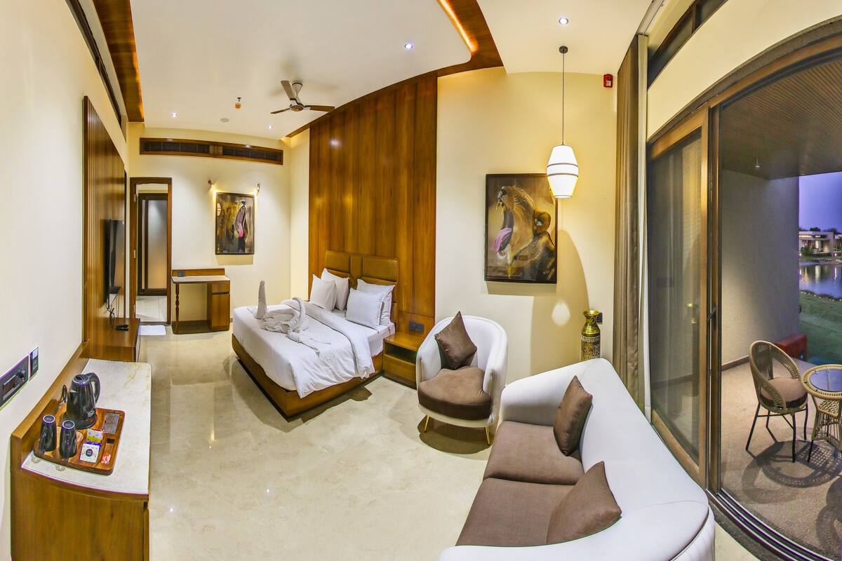 Une chambre d’hôtel moderne avec un grand lit, un canapé incurvé, un coin repas avec un plateau de produits pour le petit-déjeuner, et une vue sur une étendue d’eau par une porte vitrée.