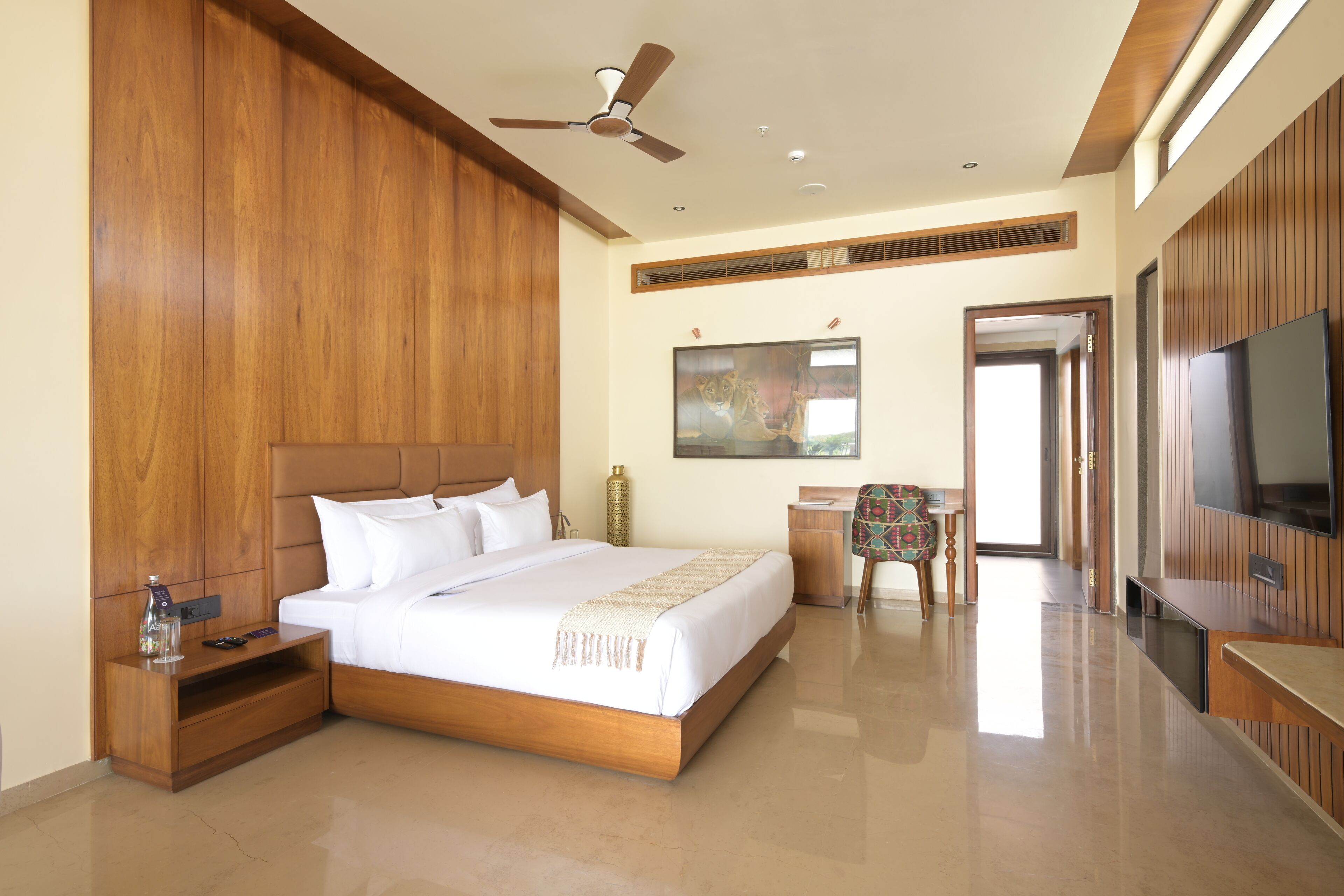 Habitación de hotel con una cama grande, paneles de madera, ventilador de techo, escritorio con silla, televisor y un cuadro en la pared.