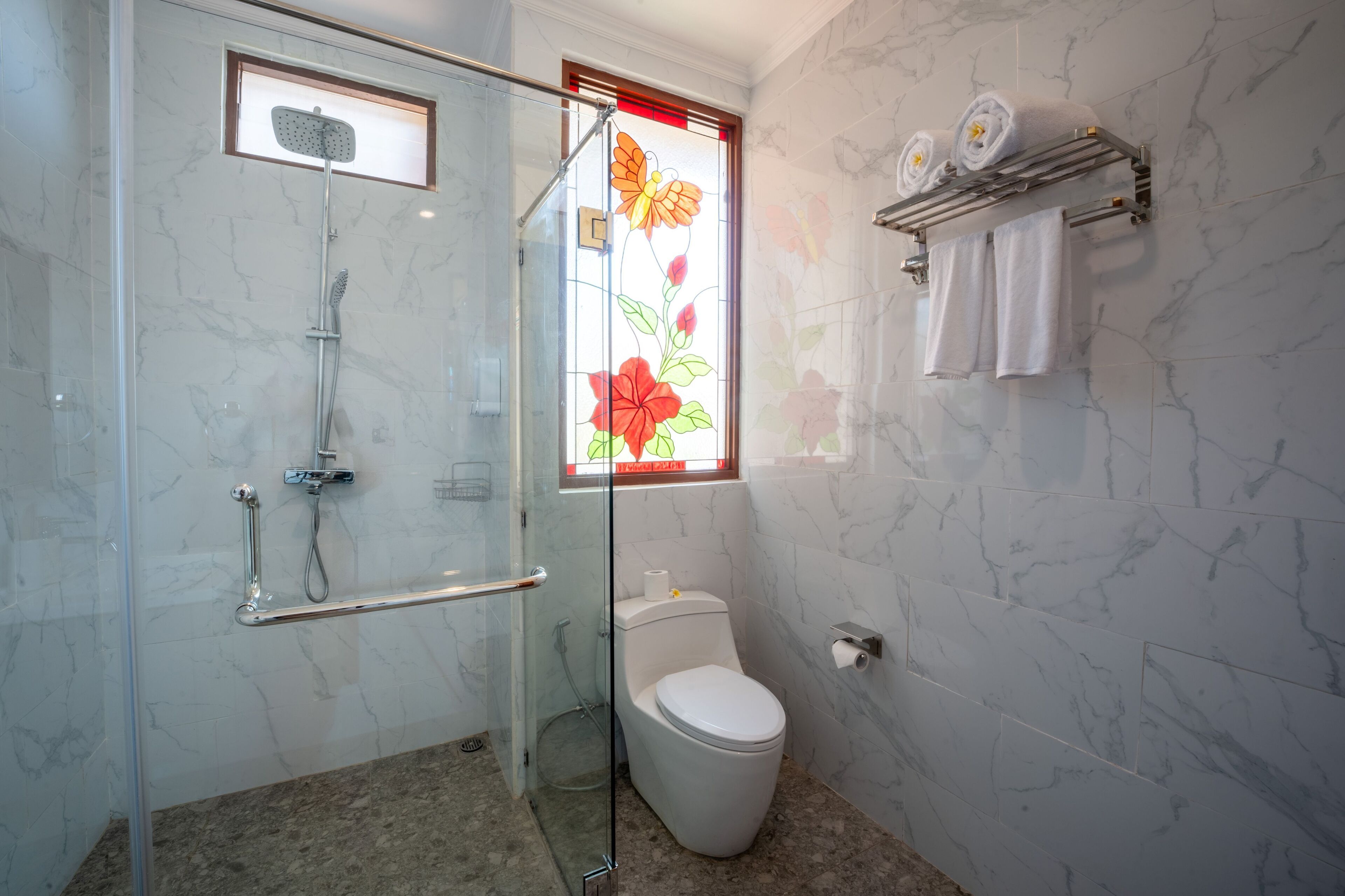 A modern bathroom with a glass-enclosed shower, a toilet, and a window with floral stained glass.