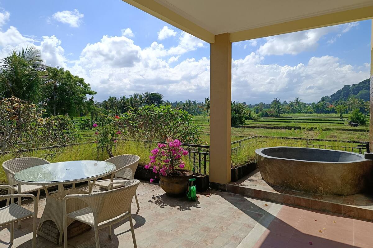 Un espacio de estar al aire libre techado con mesa y sillas, una jardinera grande con flores, y vista a un paisaje exuberante con campos escalonados.